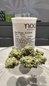 Close-up of a container labeled "Blue Dream, 3.5 Gram, Sativa - Hybrid" with cannabis buds in front. The container has detailed product information and the brand name "Noa Botanicals." Experience the renowned Blue Dream Strain like never before.