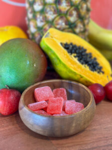 A wooden bowl with sugar-coated vegan cannabis infused gummies in front of various fruits including a mango, papaya, pineapple, banana, and apples.